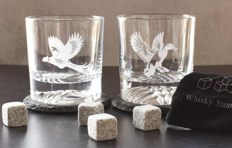 Premium laser-engraved whiskey gift set featuring two rocks glasses with game bird designs (pheasant and duck). The set includes two matching slate coasters and granite whiskey stones in a black velvet bag, presented as a high-end gift for hunters and outd