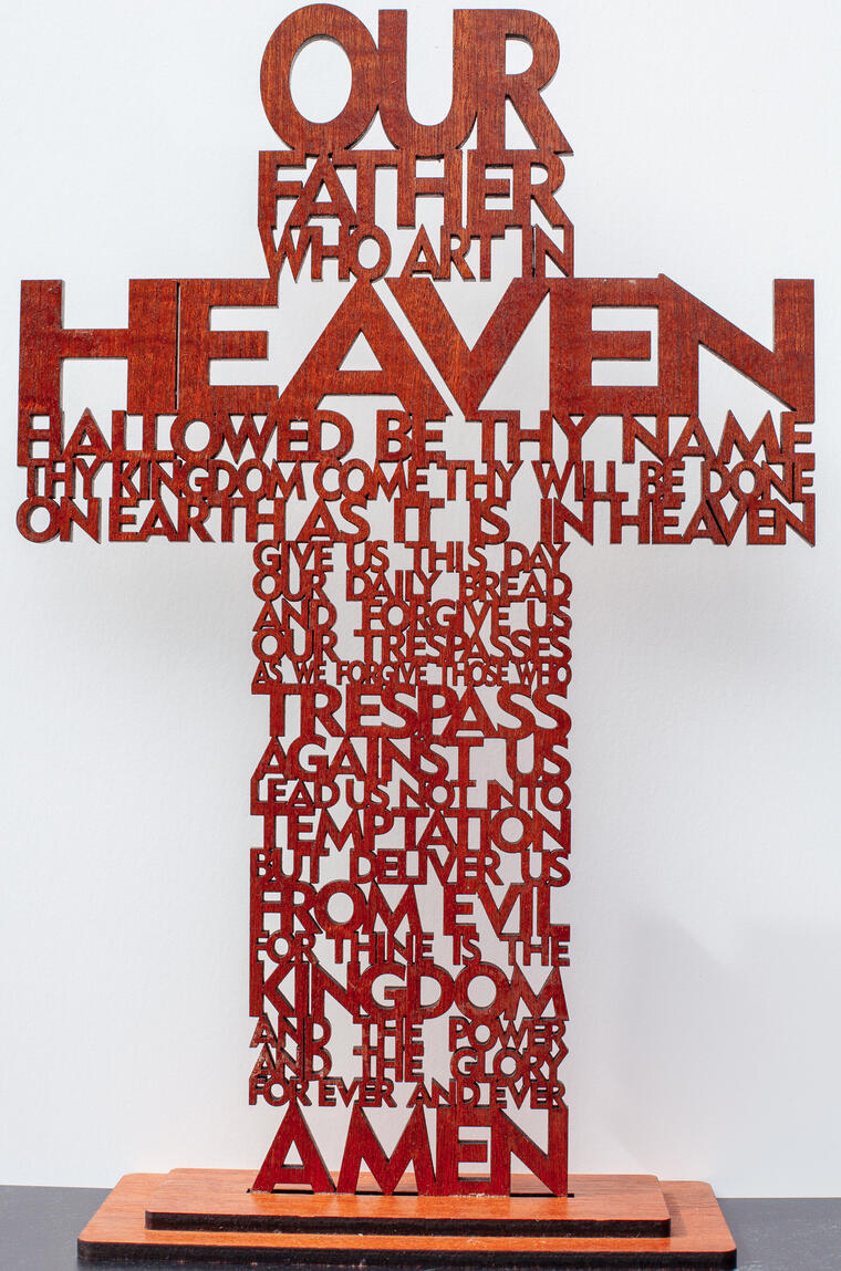 Custom laser-cut wooden cross featuring the full text of The Lord's Prayer. Crafted from 1/4 inch walnut wood with a rich dark stain, this free-standing religious decor piece showcases intricate typography and precision cutting.